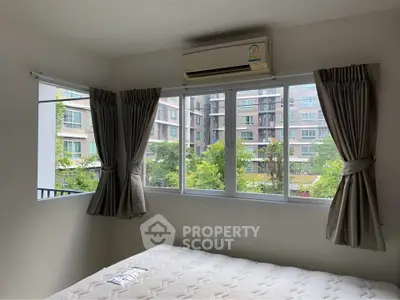Bright bedroom with large windows offering a view of modern apartment buildings and lush greenery.