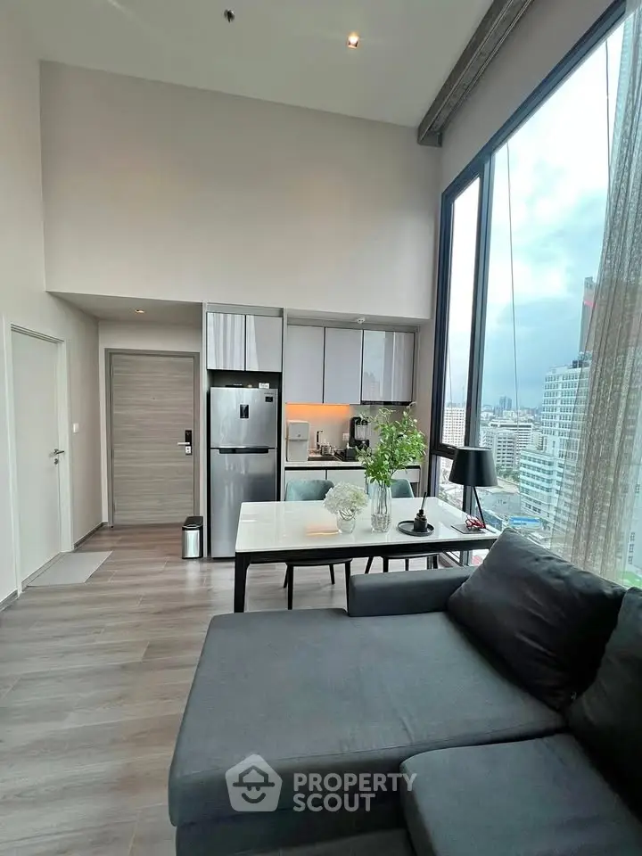 Modern living room with large windows and city view, featuring sleek furniture and open kitchen layout.