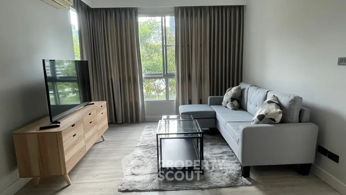 Spacious living room with modern furnishings, large windows allowing ample natural light, and a cozy grey sofa set, perfect for relaxation and entertainment.