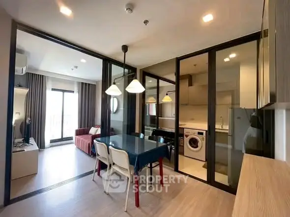 Modern open-plan living room with dining area and glass partitions, featuring a washing machine and stylish lighting.