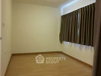 Spacious empty room with wooden flooring and large window with curtains.