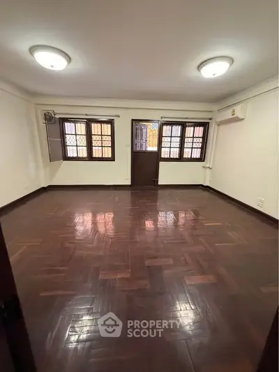 Spacious empty living room with wooden flooring and large windows in a well-lit apartment.