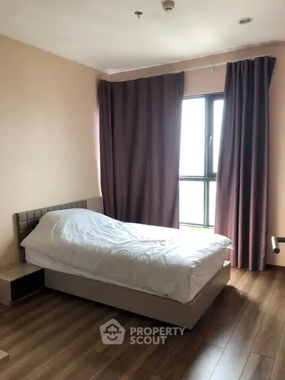Cozy bedroom with wooden flooring and large window, featuring a comfortable bed and elegant curtains.