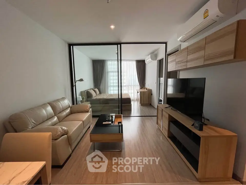 Modern open-plan living room with glass partition leading to bedroom, featuring sleek furniture and natural light.