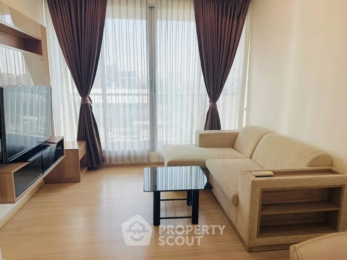 Modern living room with large windows and elegant curtains, featuring a cozy beige sofa and sleek TV setup.