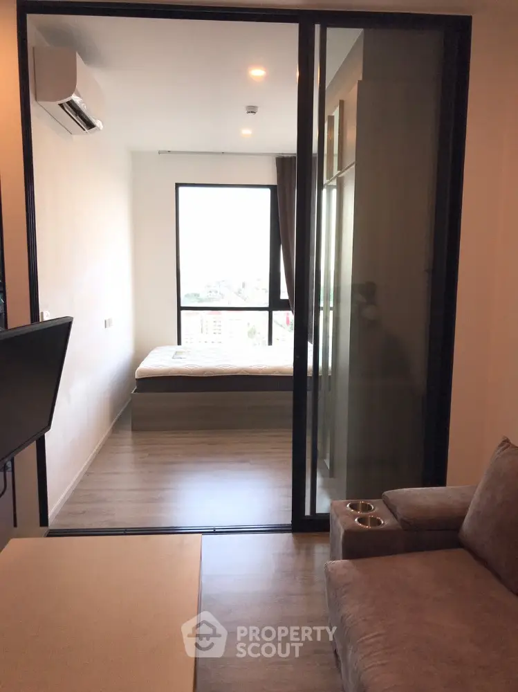 Modern apartment with sleek glass partition separating a cozy living area from a sunlit bedroom, featuring wooden flooring and contemporary furnishings.