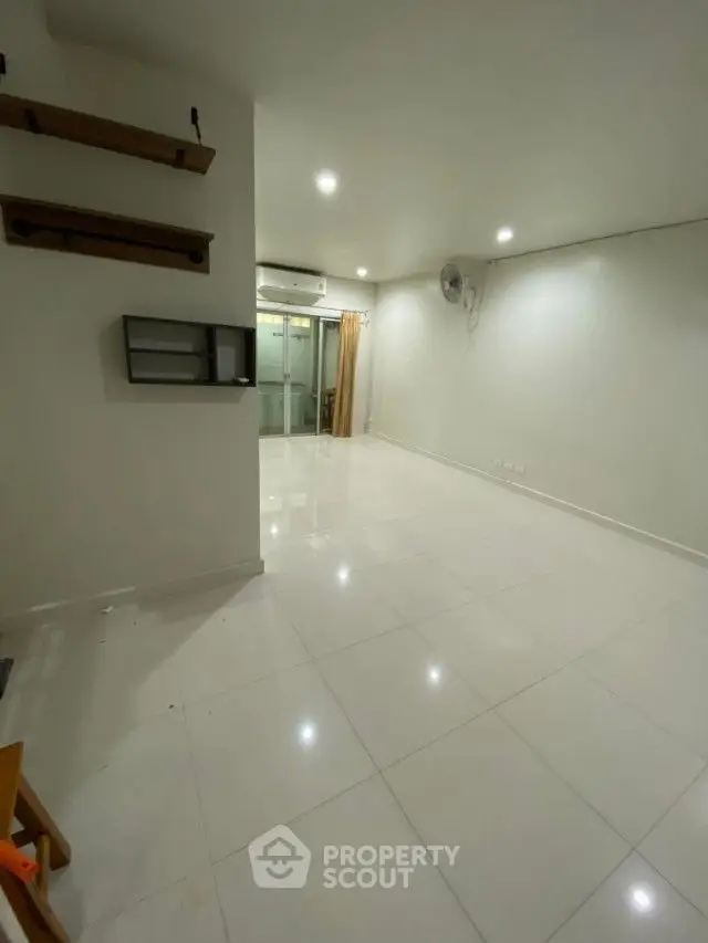Spacious and well-lit living area with glossy white tiles, modern lighting, and a sleek air conditioning unit, leading to a glass door entrance.