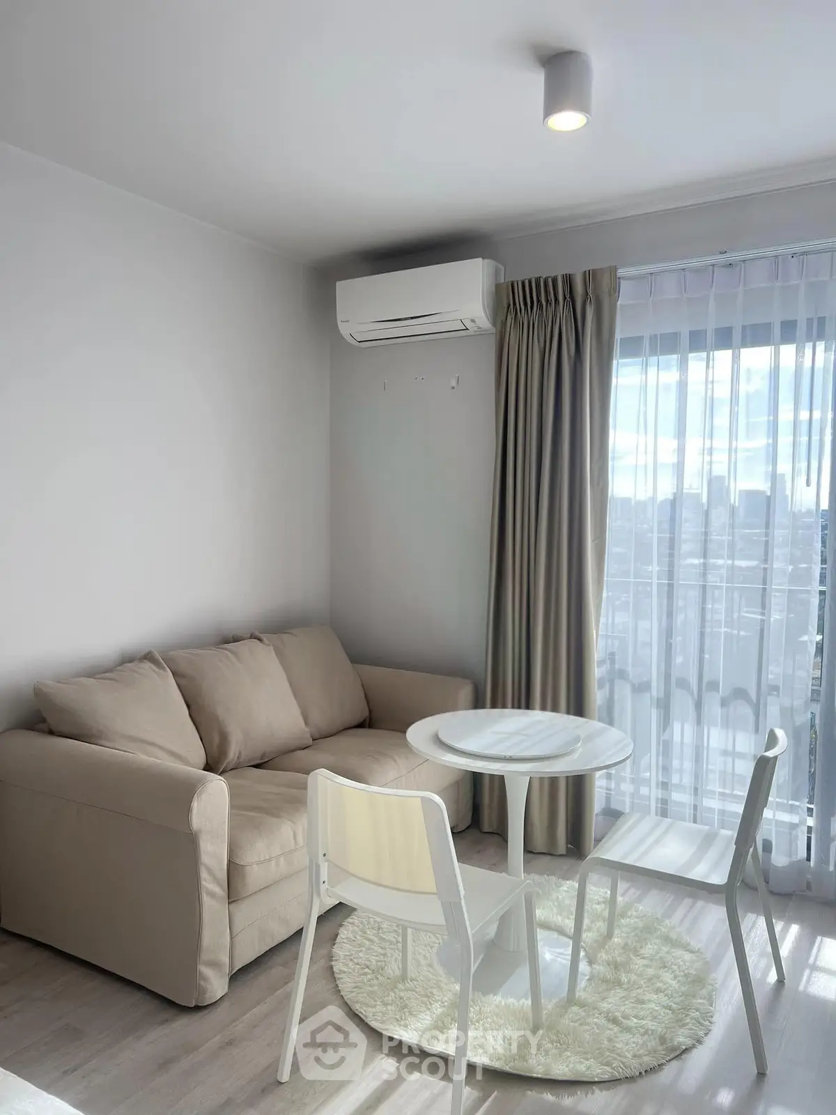 Cozy living room with beige sofa, round table, and air conditioning, featuring a bright window view.