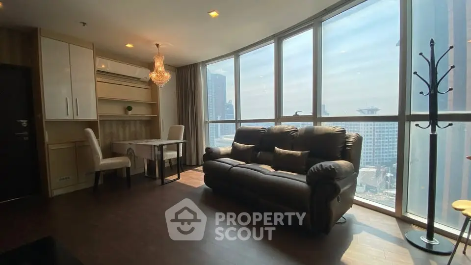 Spacious living room with large windows offering stunning city views, elegant chandelier, and cozy seating area.