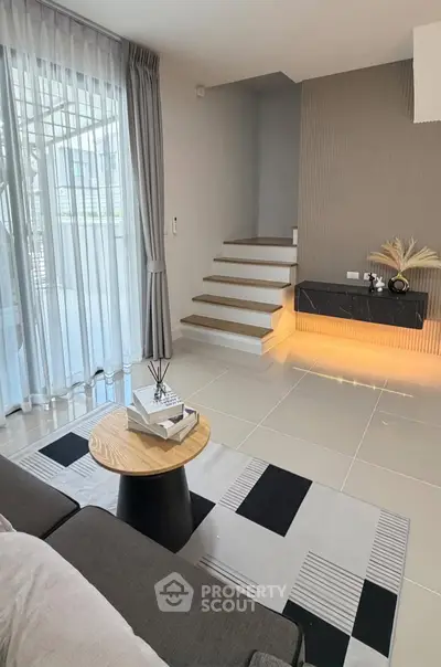 Modern living room with stylish decor and staircase, featuring large windows and elegant curtains.