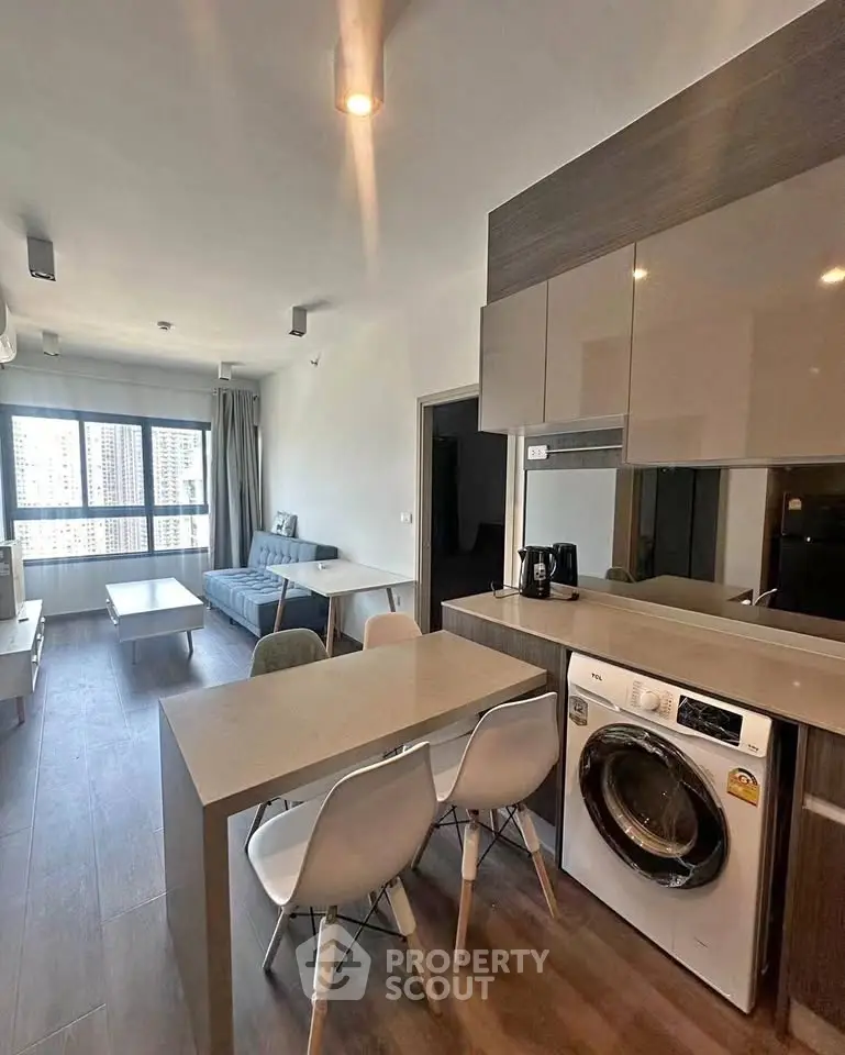 Modern open-plan living room with kitchen and washing machine, featuring sleek furniture and large window.
