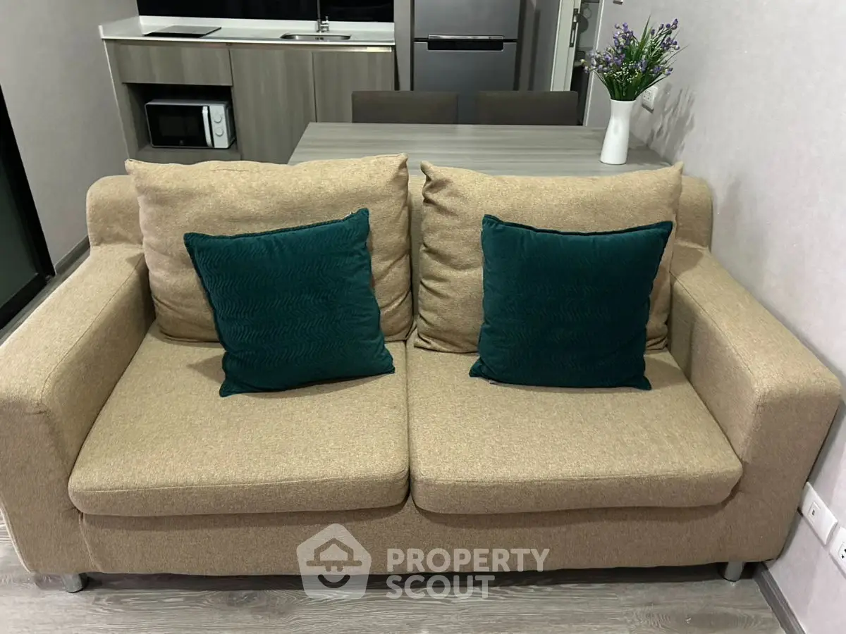 Cozy living room with beige sofa and teal cushions, modern kitchenette in background.