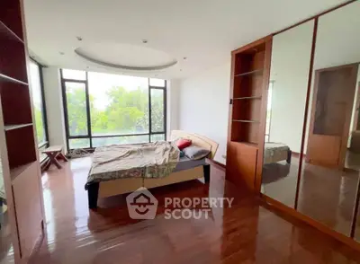 Spacious bedroom with large windows and wooden flooring, offering a serene view of greenery.