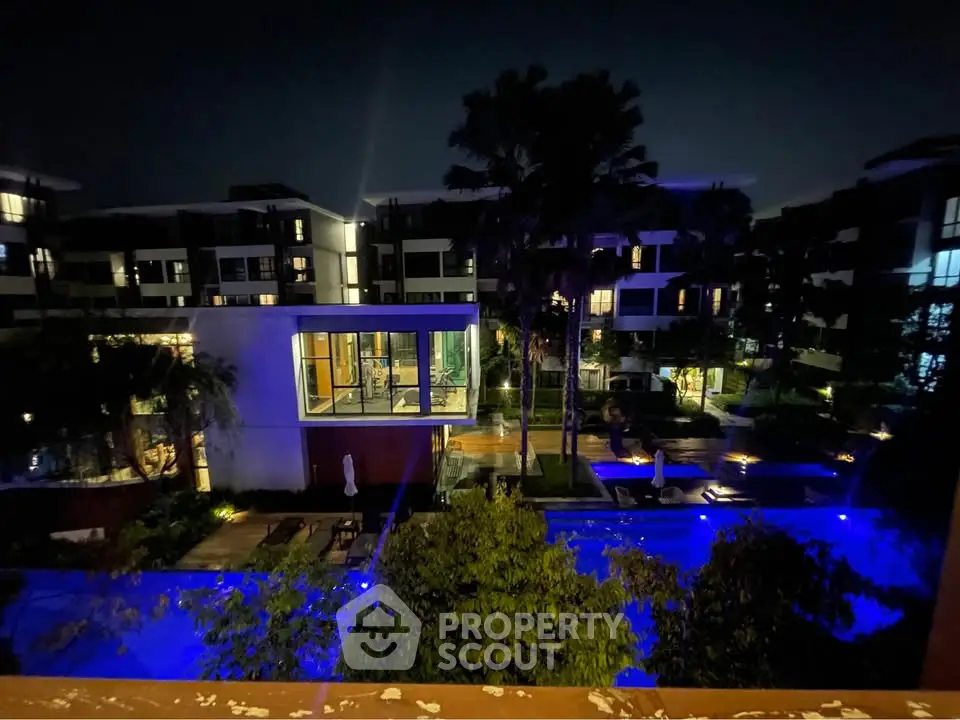 Stunning night view of modern apartment complex with illuminated pool and lush greenery.