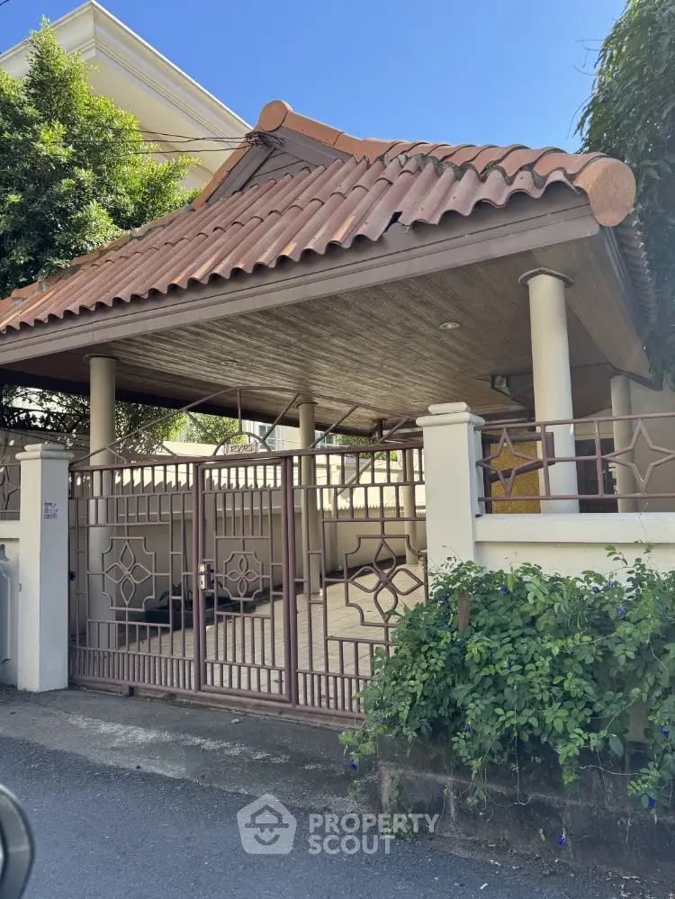 Charming residential entrance with tiled roof and gated driveway, surrounded by lush greenery.