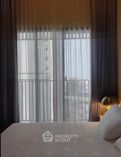 Cozy bedroom with large window and sheer curtains offering city view, perfect for relaxation.