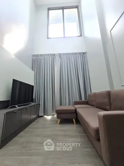 Modern living room with high ceiling and large window, featuring a cozy sofa and flat-screen TV.