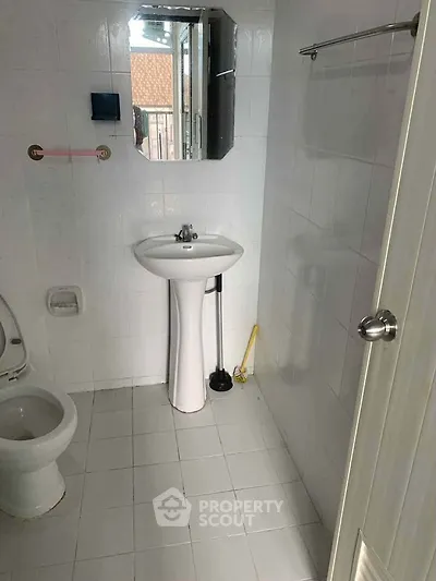 Compact bathroom with white tiles, pedestal sink, and toilet, featuring a small mirror and window for natural light.