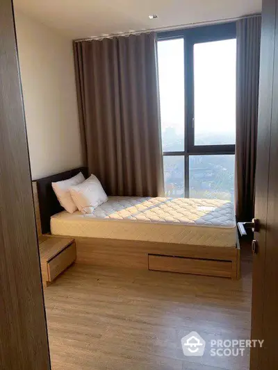 Cozy bedroom with natural light streaming through large windows, offering a serene cityscape view, complete with modern wooden furnishings.