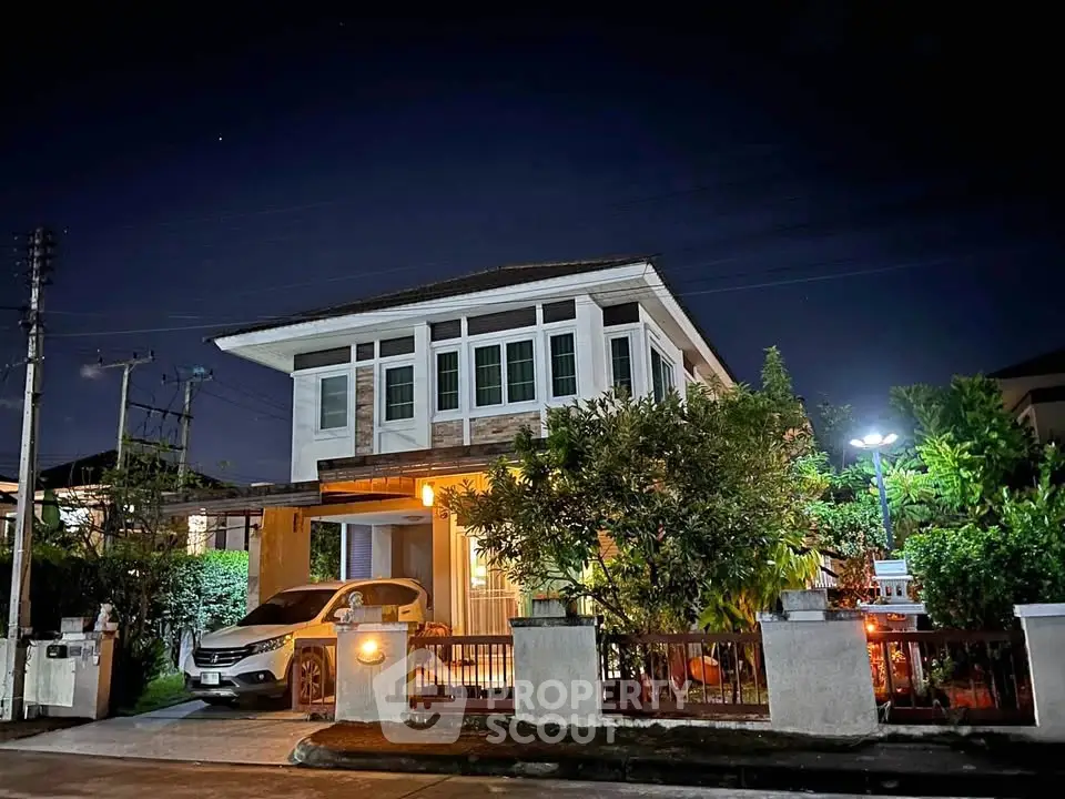 Charming two-story house with lush garden and carport, beautifully lit at night, perfect for family living.