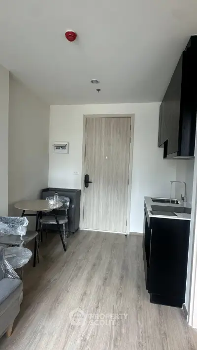 Modern apartment interior with compact kitchen and cozy dining area, featuring sleek black cabinetry and light wood flooring.
