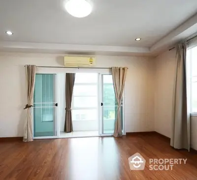 Spacious and well-lit living room with polished hardwood flooring, large windows draped with elegant curtains, and a modern air conditioning unit.