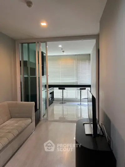 Modern living room with sleek furniture and open kitchen layout, featuring a cozy sofa and flat-screen TV.