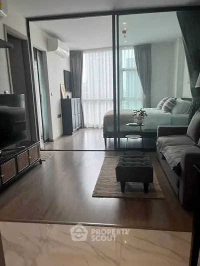 Modern open-plan living room with glass partition leading to a cozy bedroom, featuring stylish decor and natural light.