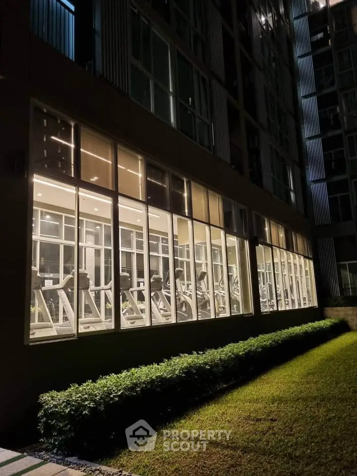 Modern gym with large windows in a residential building, showcasing fitness equipment and night lighting.