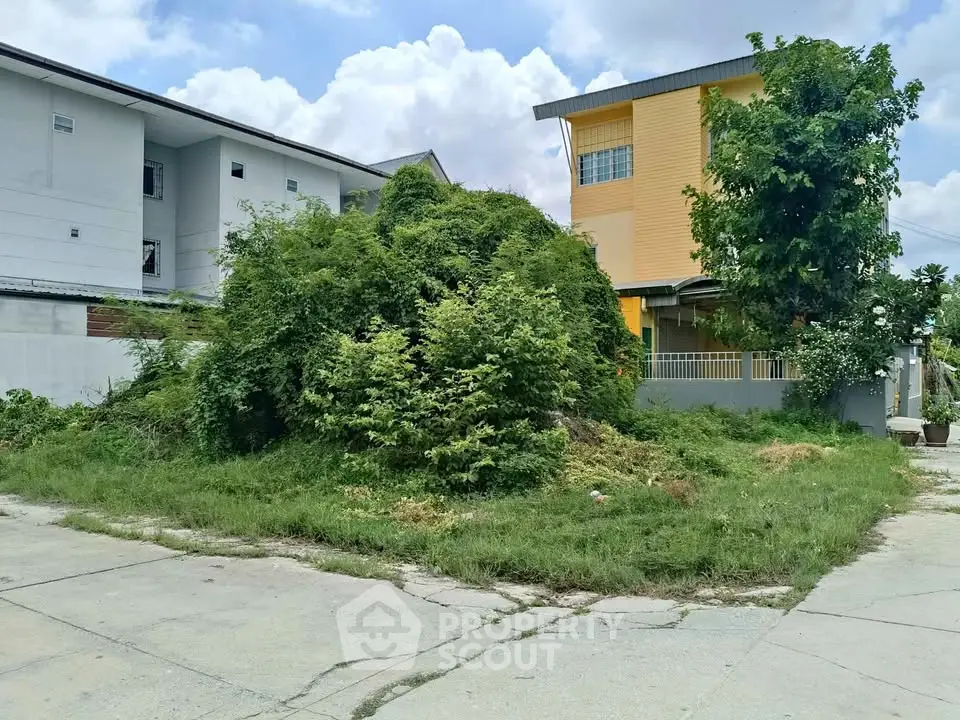 Vacant land plot with lush greenery next to residential buildings, ideal for development opportunities.