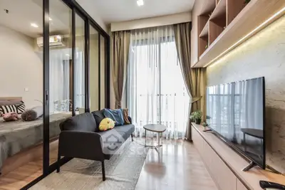Modern living room with sleek furniture and large TV, adjacent to a cozy bedroom separated by glass doors.
