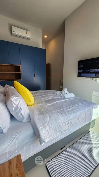 Modern bedroom with blue wardrobe and wall-mounted TV, featuring a cozy bed and bright yellow pillow.