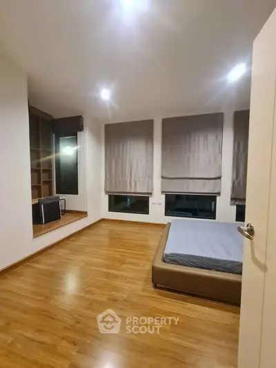 Spacious bedroom with wooden flooring and large windows, featuring a cozy bed and built-in shelving.