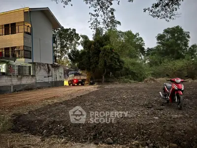 Vacant land plot with nearby residential building and parked motorcycle, ideal for development.