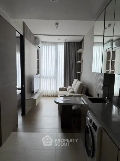 Modern compact living room with integrated kitchen and washing machine, featuring sleek design and natural light.