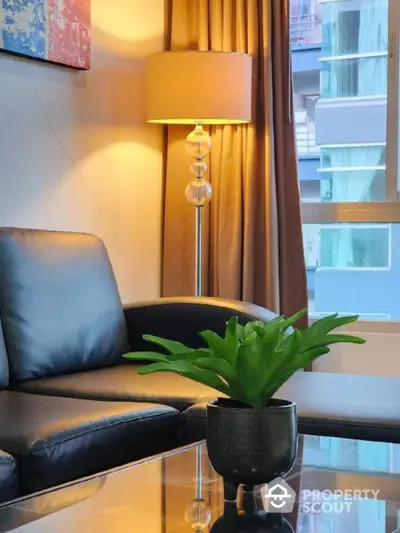 Elegant living space with modern leather sofa, stylish floor lamp, and vibrant green plant adding a touch of nature, set against warm drapery and city views.