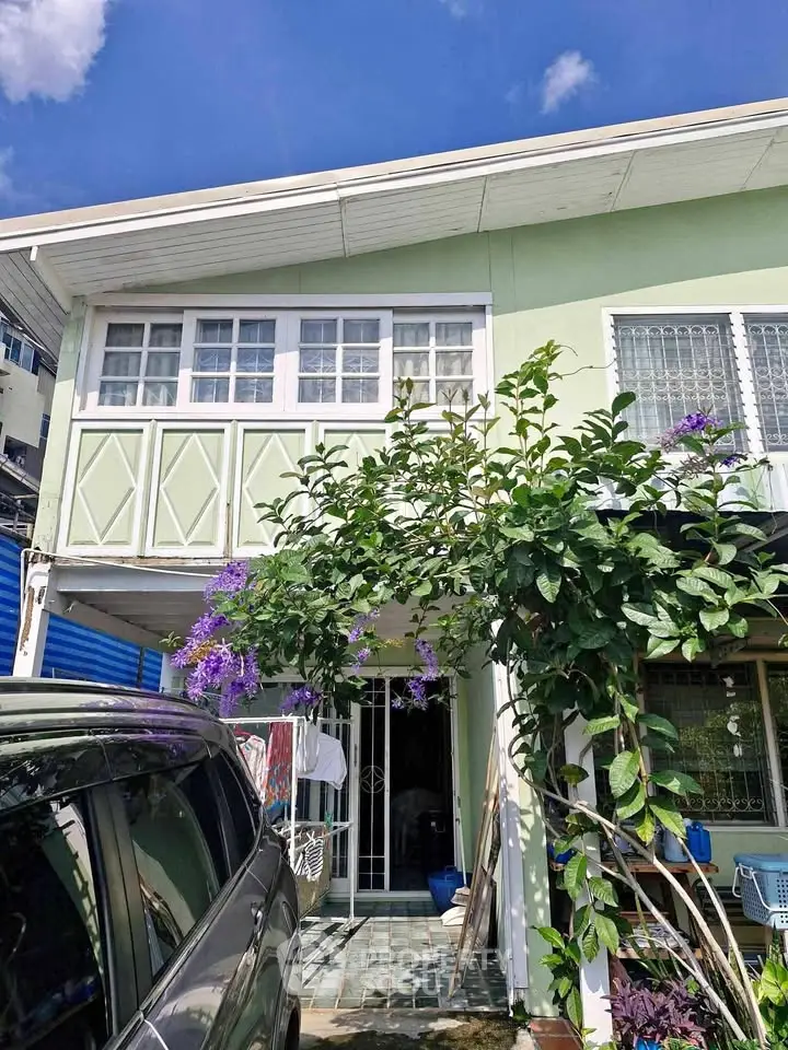 Charming two-story green house with lush garden and car parked in driveway under clear blue sky.