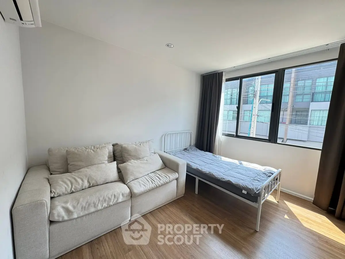 Bright studio apartment with cozy sofa and bed, featuring large windows and wooden flooring.