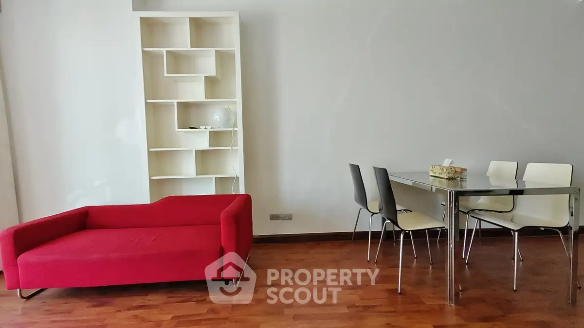 Modern living room with red sofa, glass dining table, and sleek shelving unit on wooden flooring.