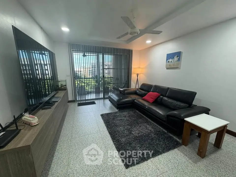 Spacious living room with modern decor and large TV, featuring a cozy black leather sofa and balcony access.