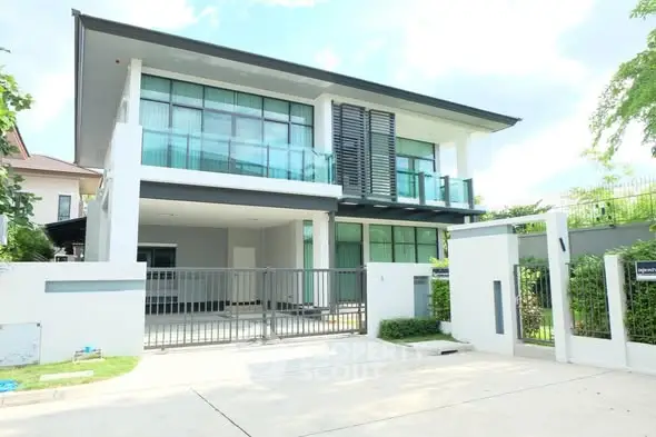 Modern two-story house with sleek design and large windows, featuring a gated entrance and lush greenery.