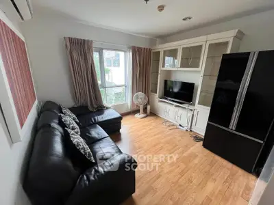 Cozy living room with black leather sofa, TV unit, and large fridge, featuring wooden flooring and natural light.