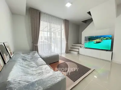 Modern living room with sleek furniture and large TV, featuring elegant staircase and natural light.