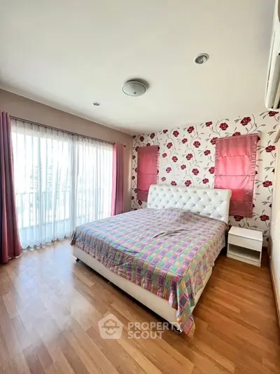 Bright bedroom with floral wallpaper and large window, featuring a cozy bed and wooden flooring.