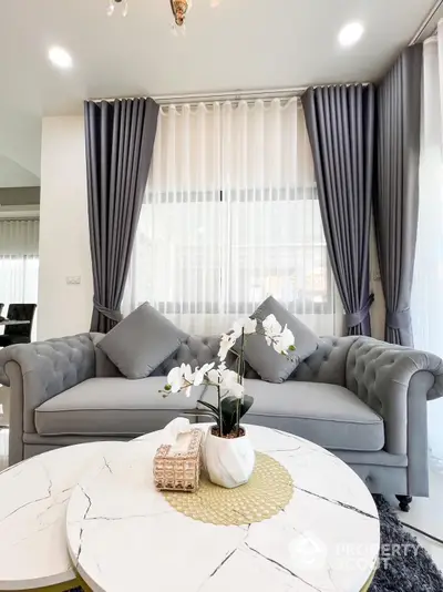 Elegant living room with luxurious grey tufted sofa, chic marble coffee table, and sophisticated drapery, bathed in natural light.