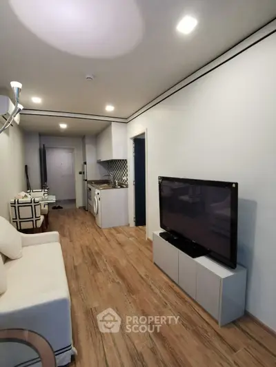 Modern living room with open kitchen and wooden flooring, featuring a cozy sofa and flat-screen TV.