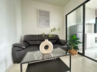 Modern living room with sleek black leather sofa and glass coffee table, adjacent to a bright kitchen area.