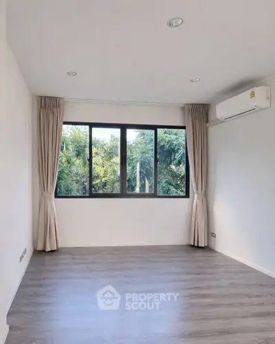 Spacious empty room with large window and air conditioning, offering serene garden views.