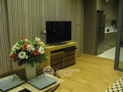 Modern living room with stylish decor and open kitchen layout, featuring a sleek TV setup and elegant floral arrangement.