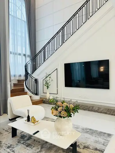 Luxurious living room with elegant staircase and modern decor, featuring a large flat-screen TV and stylish furniture.